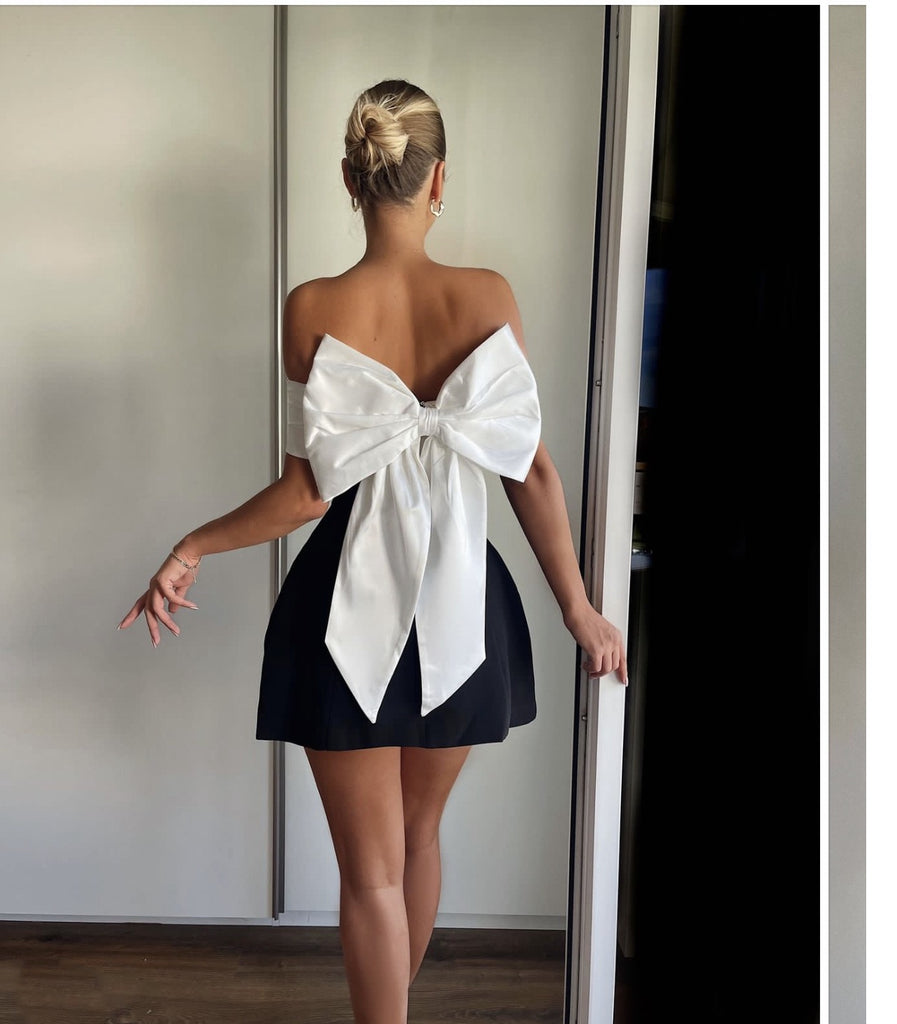 Woman wearing the Lila Bow-Back Mini Dress featuring an oversized satin bow, off-shoulder neckline, and a chic black and white design.