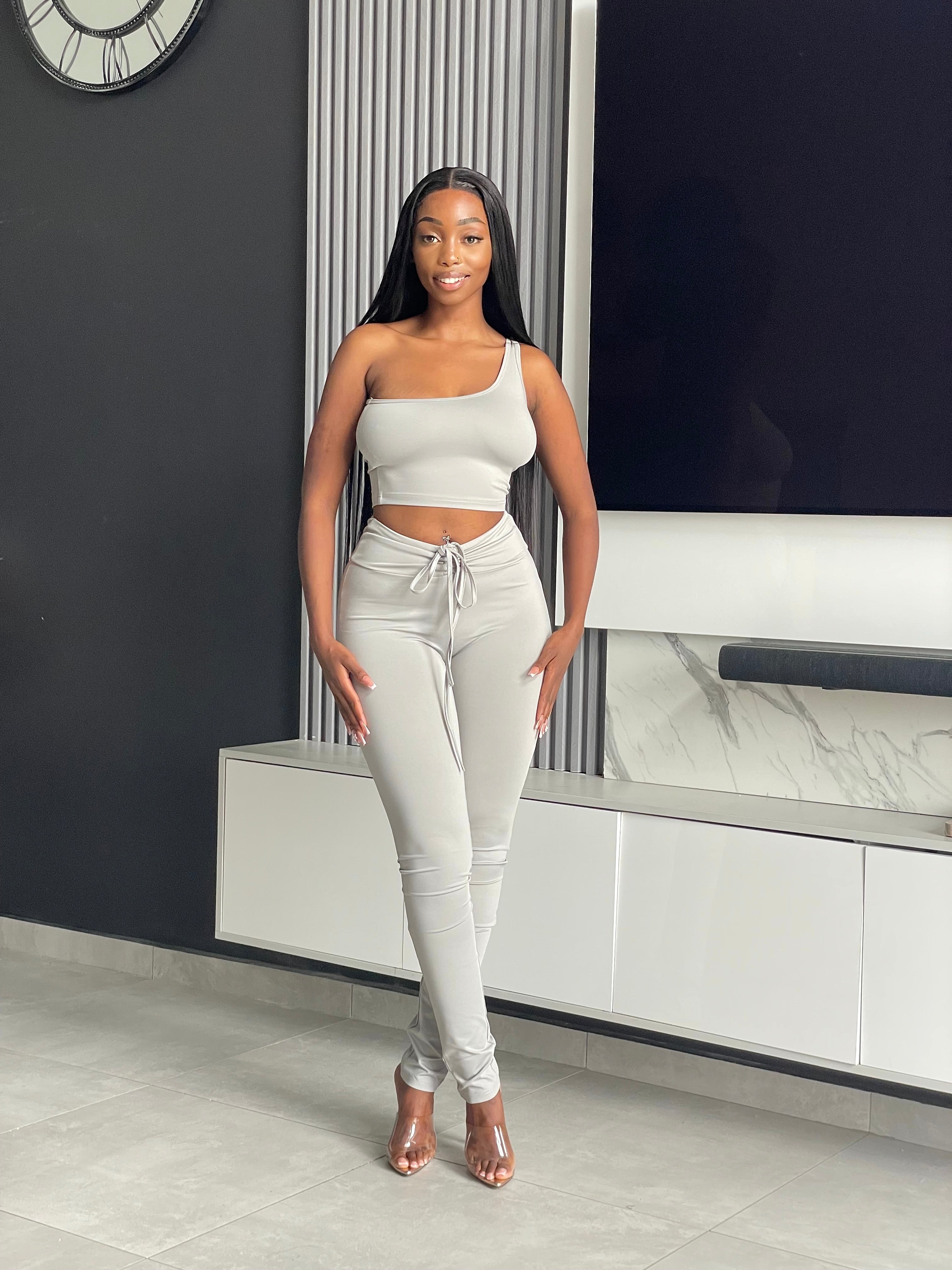 Model wearing the Ama Pant Set in light grey, featuring a one-shoulder crop top and jogger-style pants with drawstring.