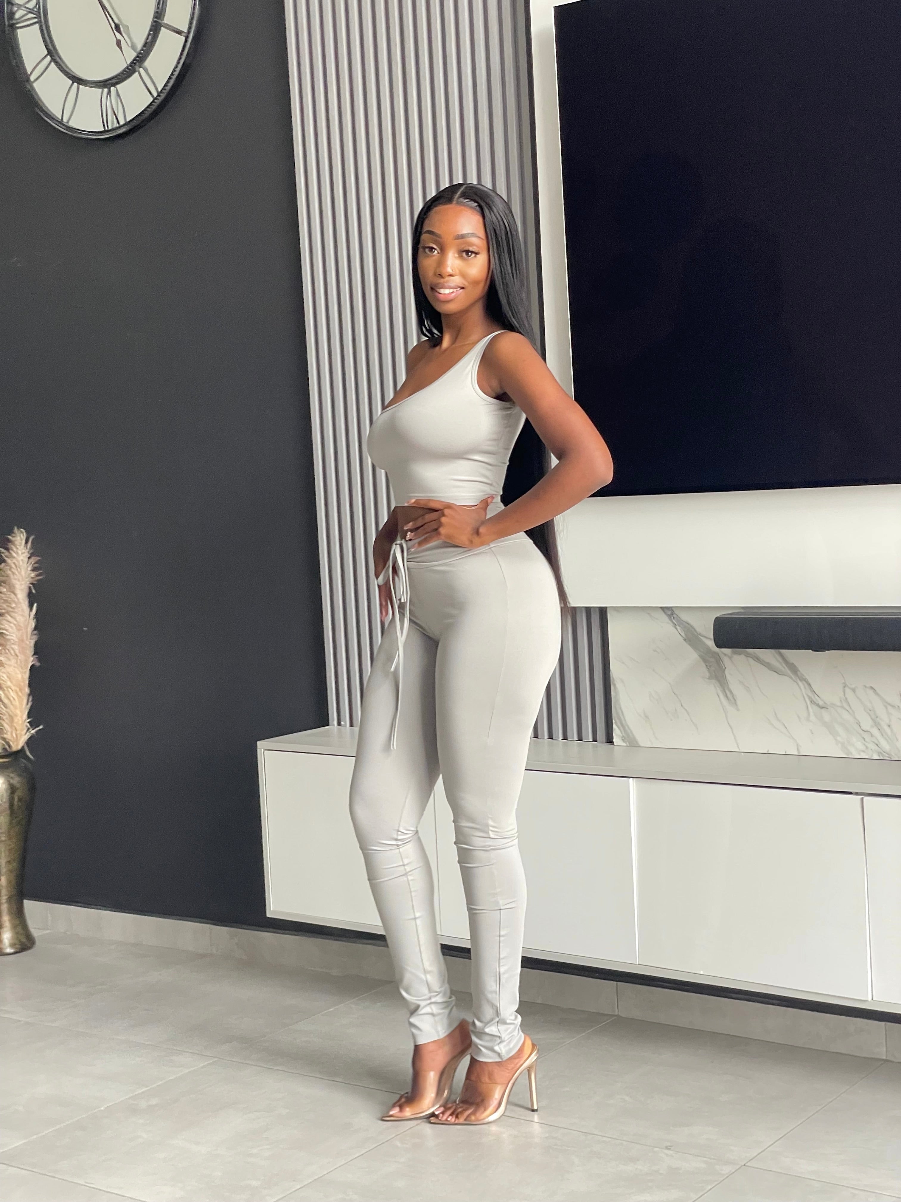 Model wearing the Ama Pant Set in light grey, featuring a one-shoulder crop top and jogger-style pants with drawstring.