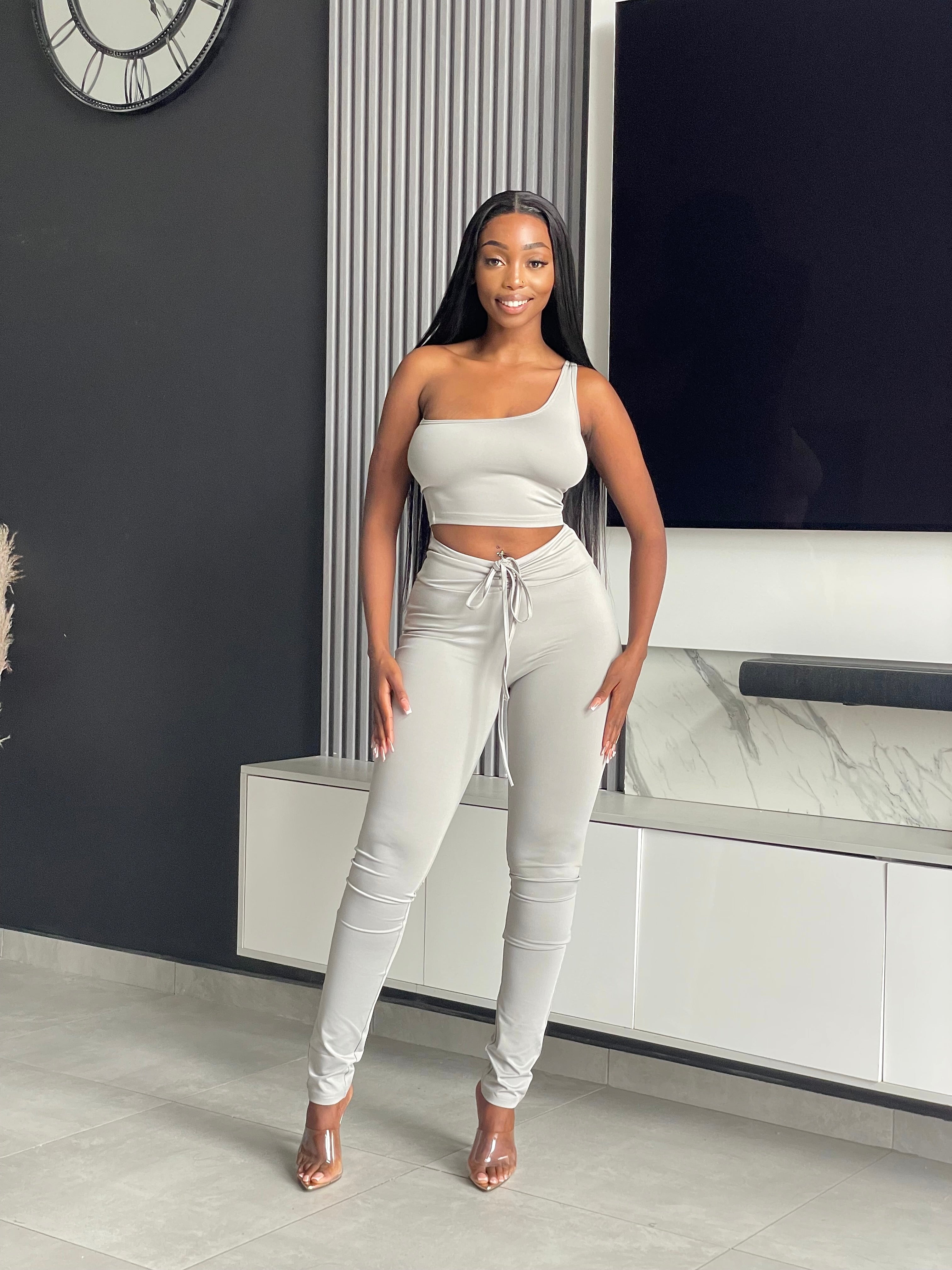 Model wearing the Ama Pant Set in light grey, featuring a one-shoulder crop top and jogger-style pants with drawstring.