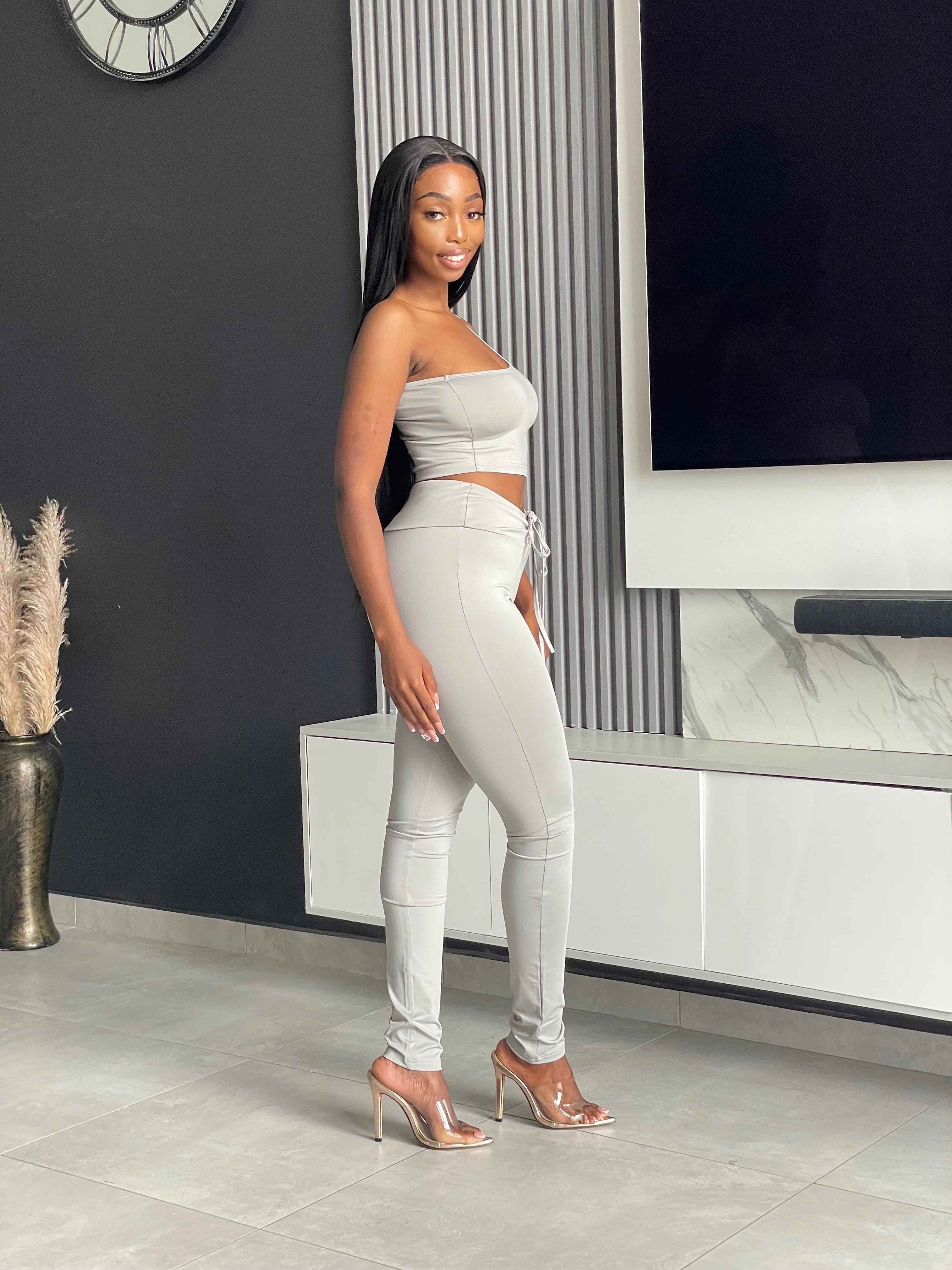 Model wearing the Ama Pant Set in light grey, featuring a one-shoulder crop top and jogger-style pants with drawstring.